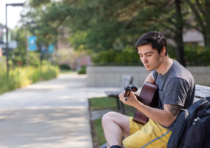 Student playing guitar outside on a bench.